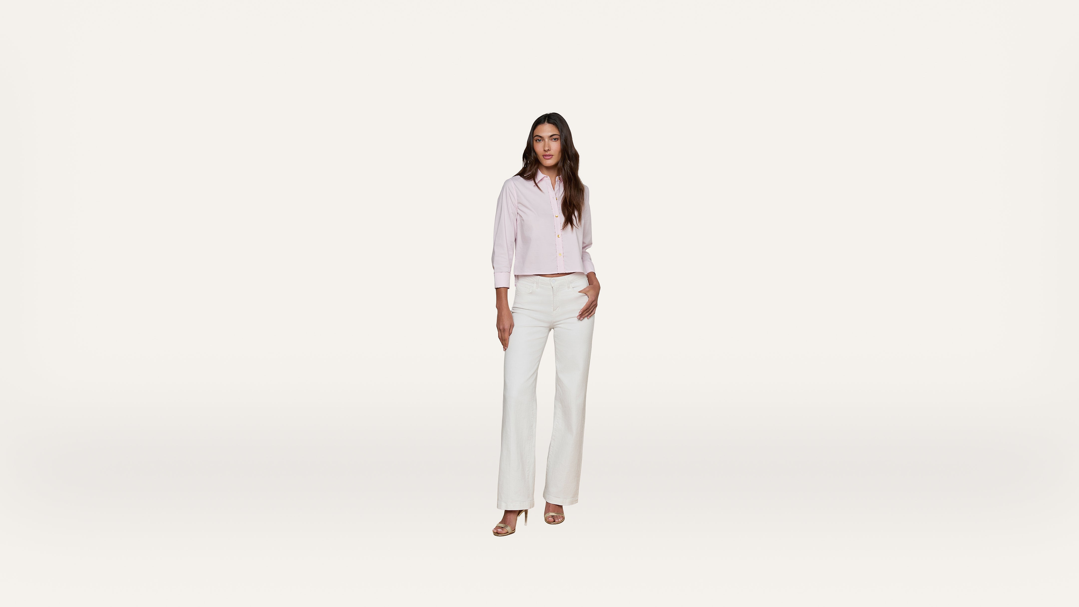 A woman stands against a plain white background, wearing a light pink long-sleeve button-up shirt, white wide-leg pants, and sandals.