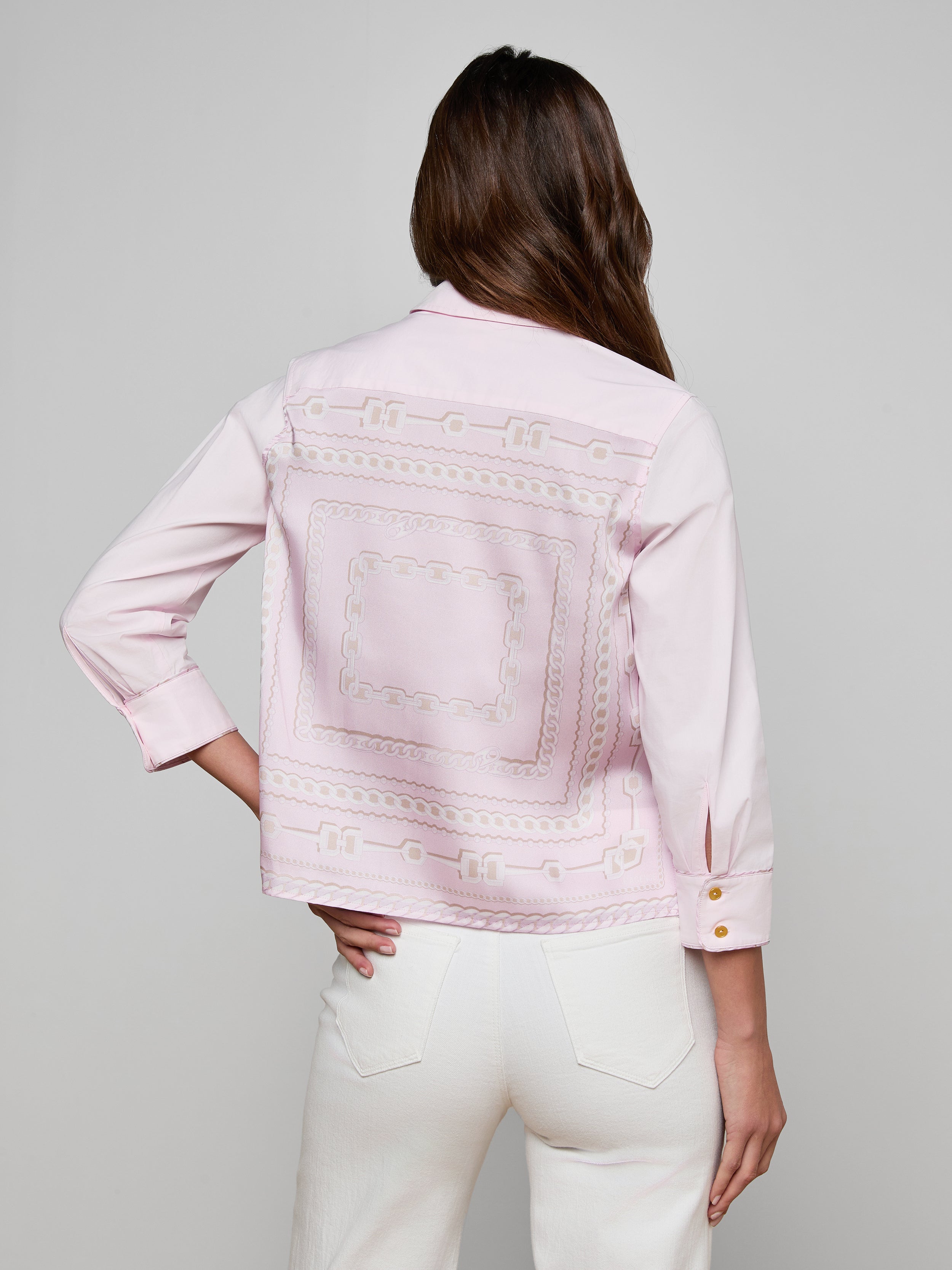 A woman with long brown hair, standing with one hand in her pocket and facing away from the camera against a plain background, wears the Fawn Shirt—a cropped pale pink top featuring embroidered patterns on the back—paired with white pants.