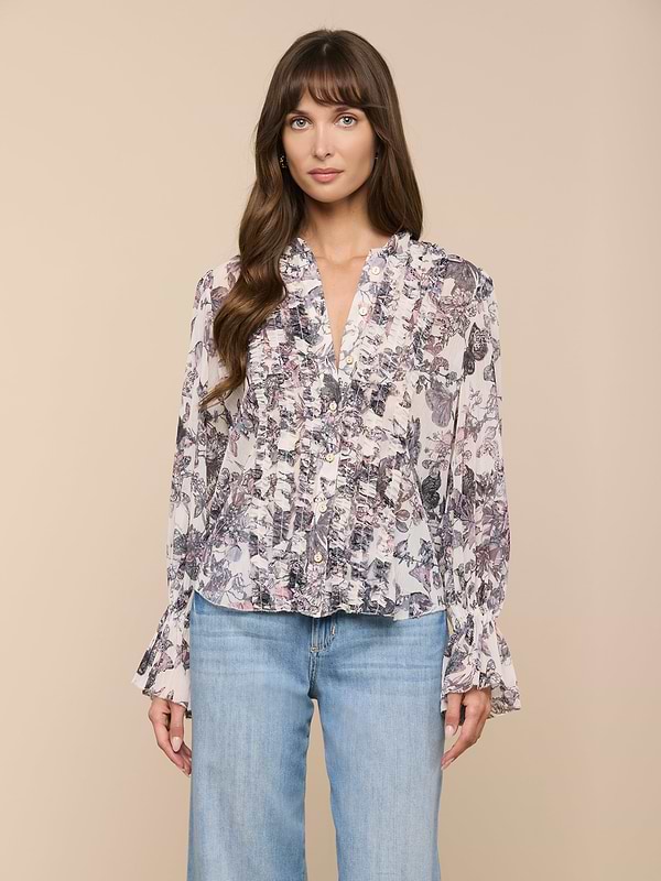 A woman is wearing the Eunice Blouse, a long-sleeve butterfly print top with ruffled cuff details, paired with light blue jeans and standing against a plain, light background.