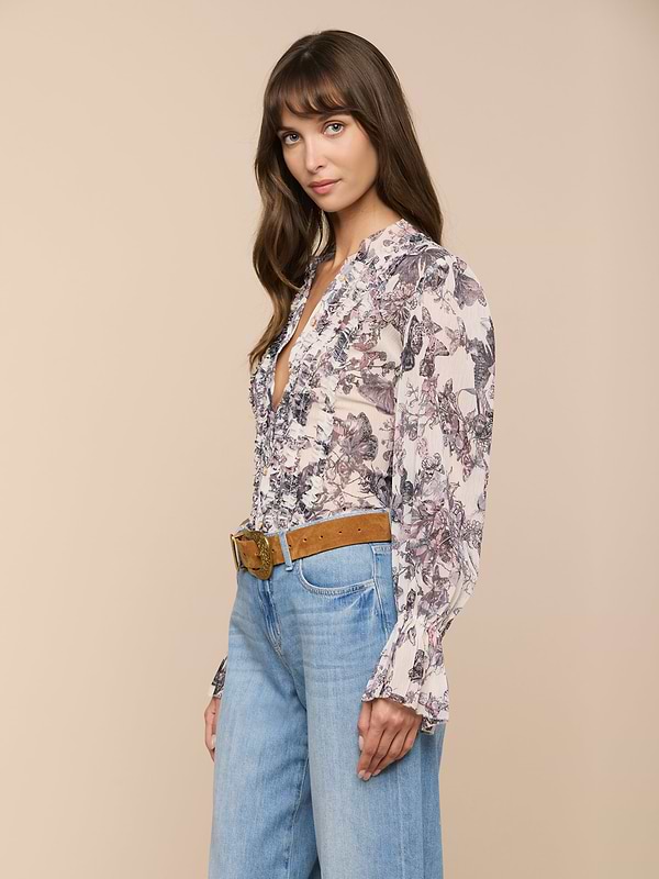 Woman with long brown hair stands against a plain background, wearing the Eunice Blouse—a butterfly print top—paired with blue high-waisted jeans and a tan belt. She looks slightly to her left with a relaxed expression, her hands by her sides.
