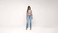 A woman stands against a plain white background wearing sunglasses, a floral blouse, blue high-waisted jeans, a brown belt, and open-toed heels.