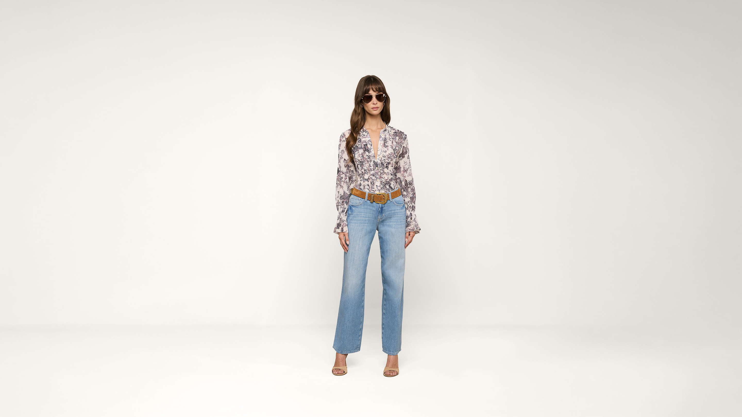 A woman stands against a plain white background wearing sunglasses, a floral blouse, blue high-waisted jeans, a brown belt, and open-toed heels.