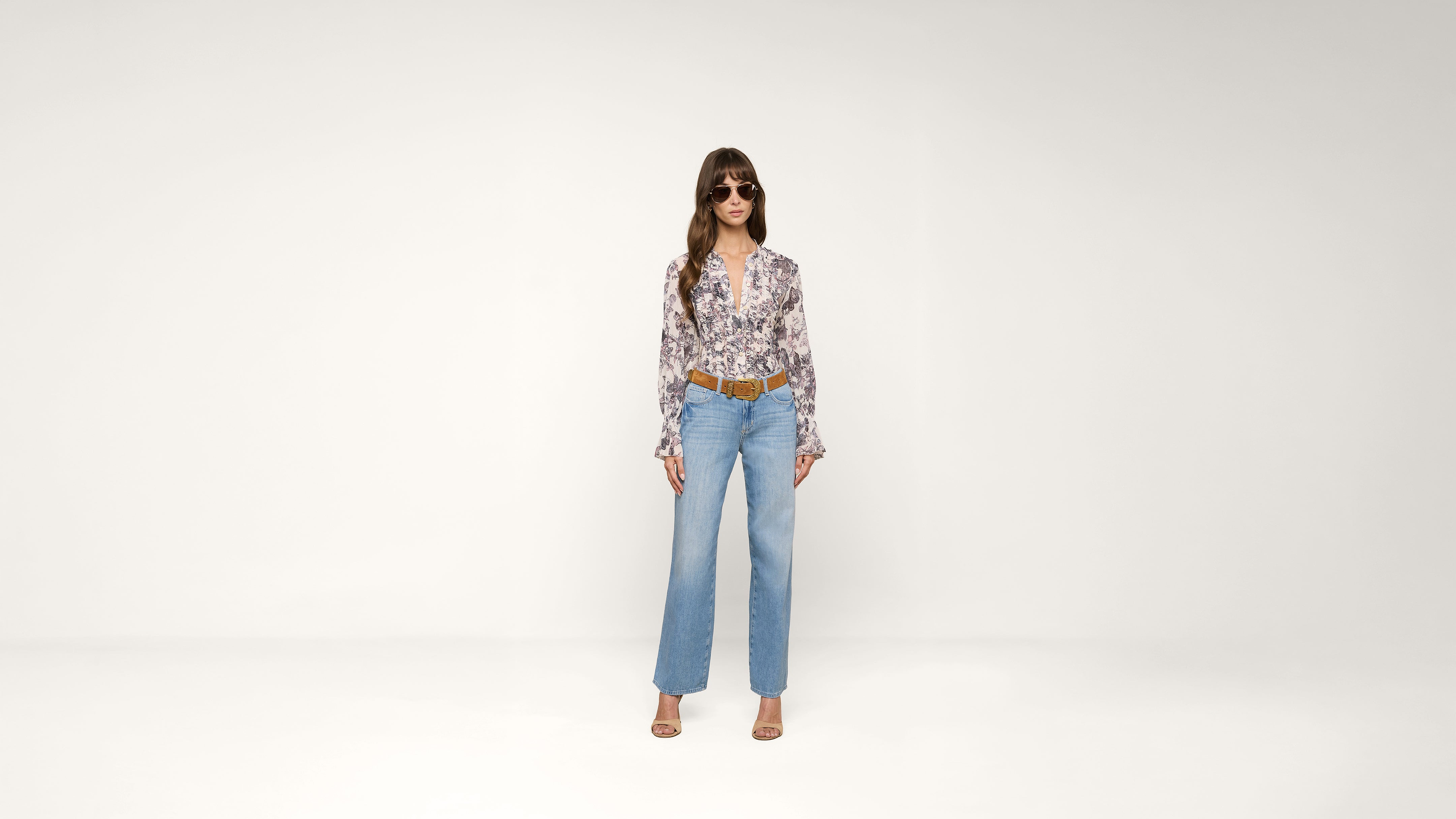A woman stands against a plain white background wearing sunglasses, a floral blouse, blue high-waisted jeans, a brown belt, and open-toed heels.