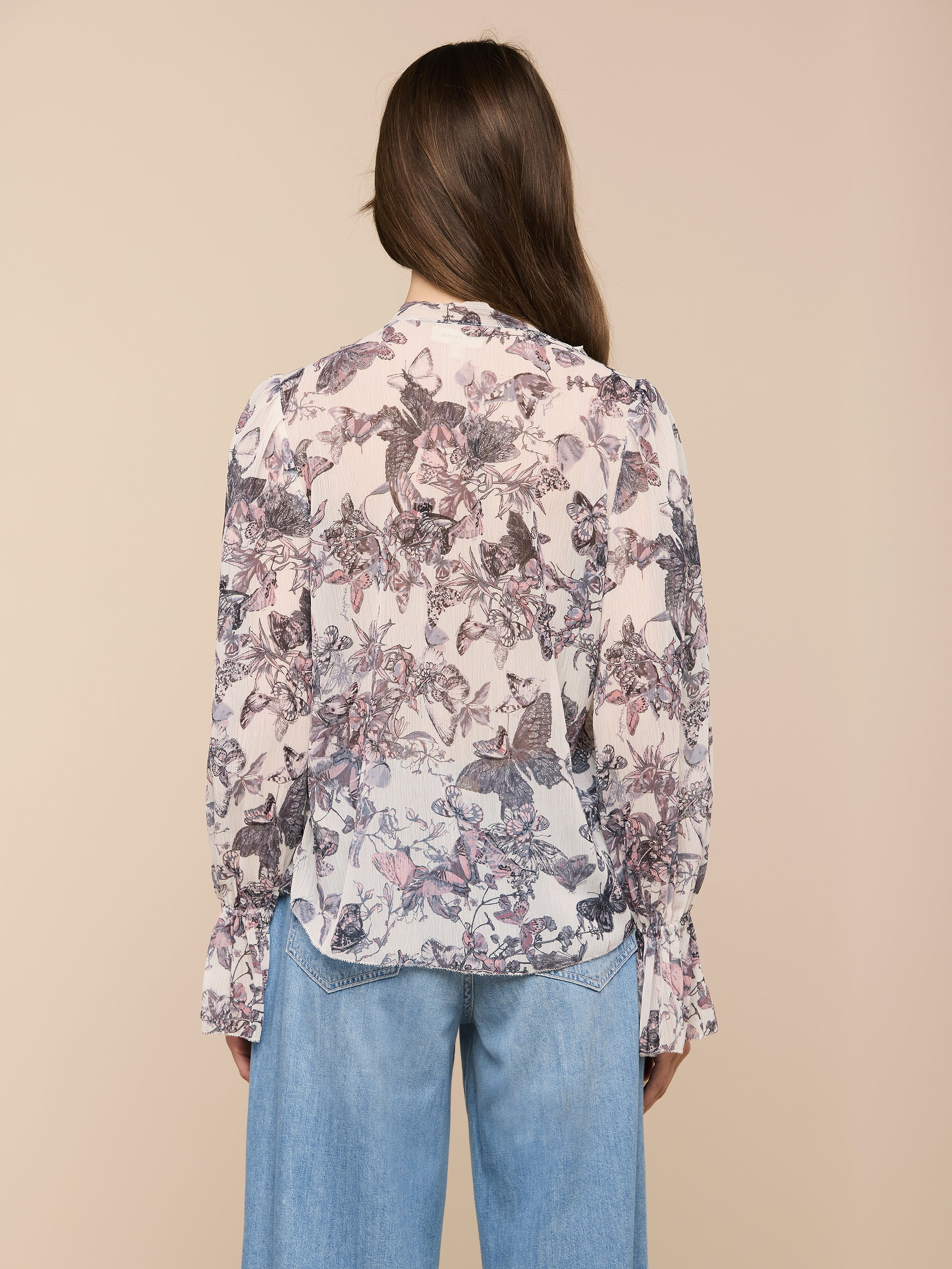 A woman with long brown hair is shown from the back against a plain light background, wearing the Eunice Blouse featuring a butterfly print and paired with light blue jeans.