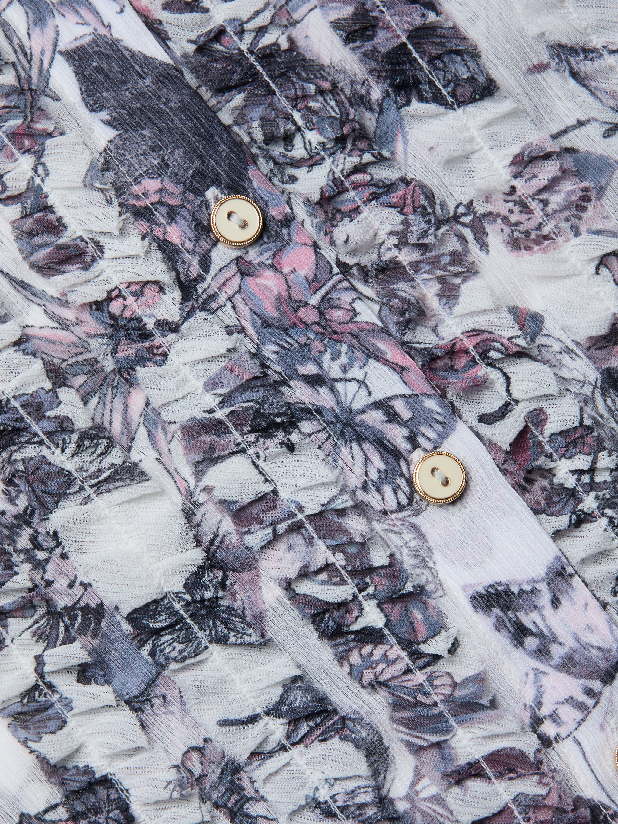 Close-up of the Eunice Blouse fabric featuring a pink, purple, and gray floral pattern, with two beige buttons bordered by gold rims sewn onto it. The textured, slightly sheer material showcases visible stitching lines—details reminiscent of those found on ruffled cuffs or butterfly print blouses.