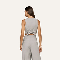 Woman facing away, wearing the Arwen Vest in light gray—a lightweight wool blend cropped vest with a knotted detail at the back—paired with matching high-waisted pants. She has long brown hair and is accessorized with earrings.