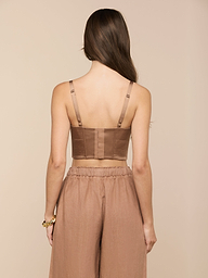 A woman with long brown hair faces away, wearing the Nysa Bustier Top—a brown cropped top with adjustable straps—and loose brown pants. She has a gold bracelet on her left wrist. The background is plain and light.
