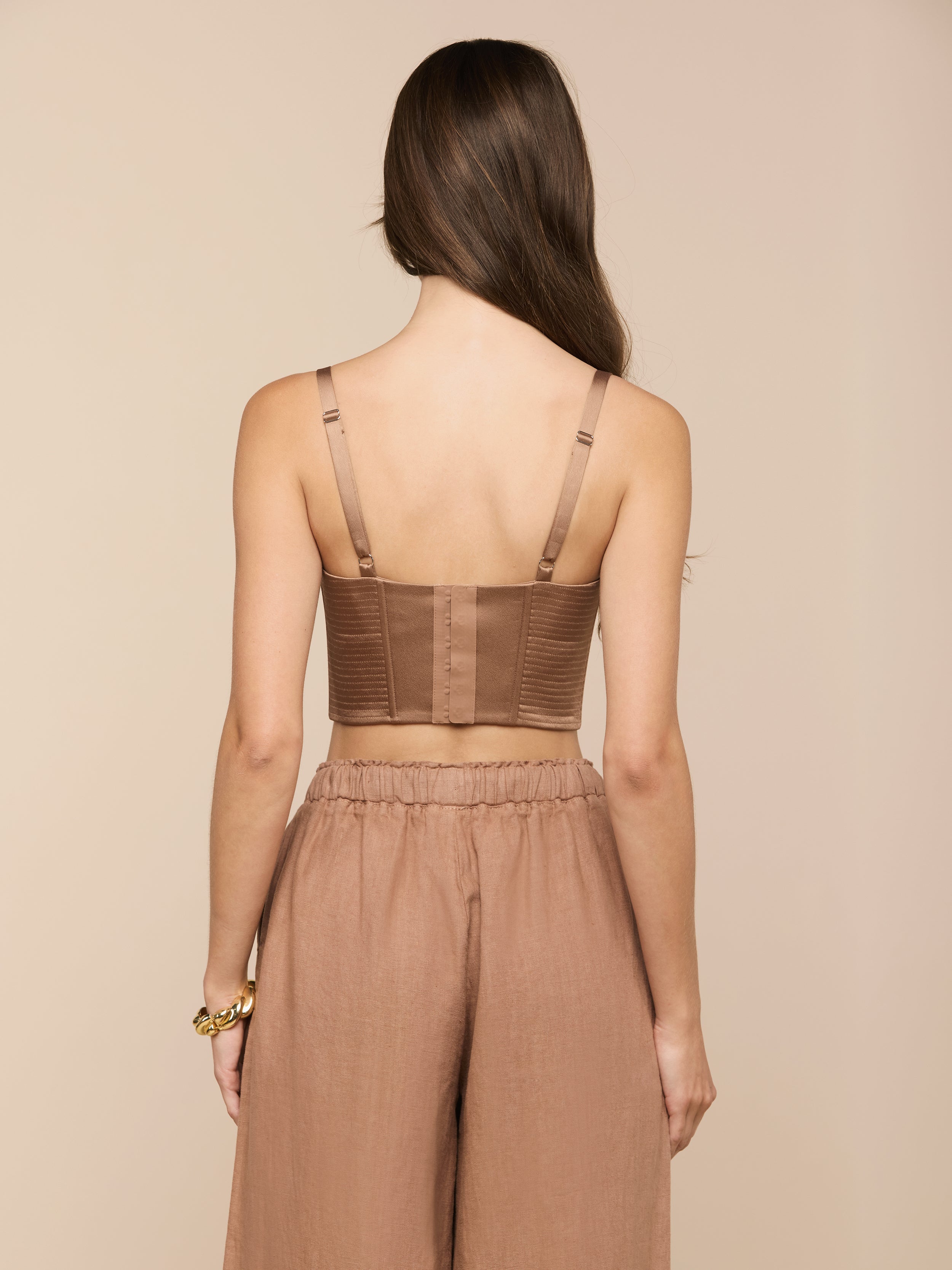 A woman with long brown hair faces away, wearing the Nysa Bustier Top—a brown cropped top with adjustable straps—and loose brown pants. She has a gold bracelet on her left wrist. The background is plain and light.