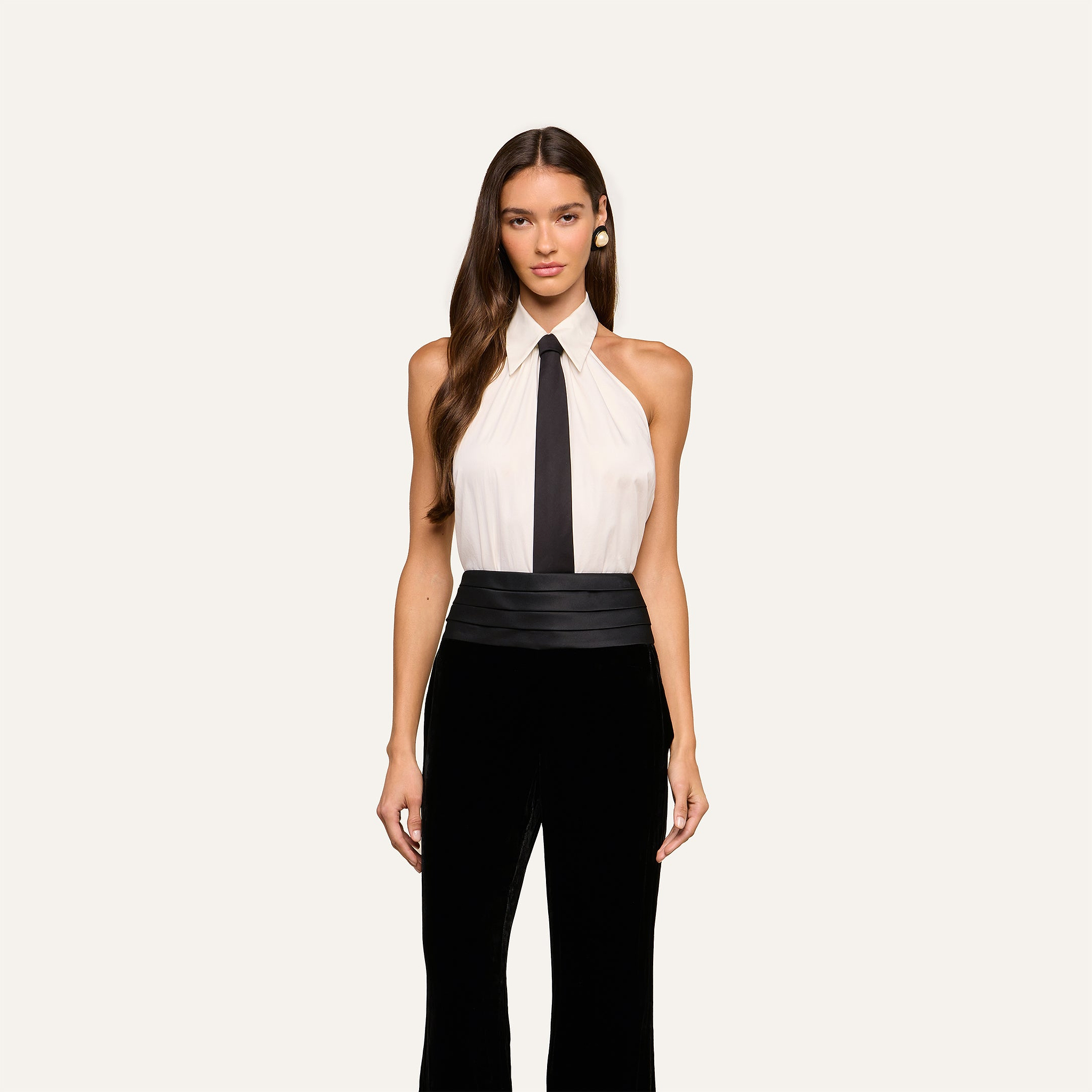 A woman with long brown hair wears the Wren Neck-Tie Top— a sleeveless, halter black and white button-down piece featuring a black tie detail—paired with wide-leg pants, standing against a plain light background.