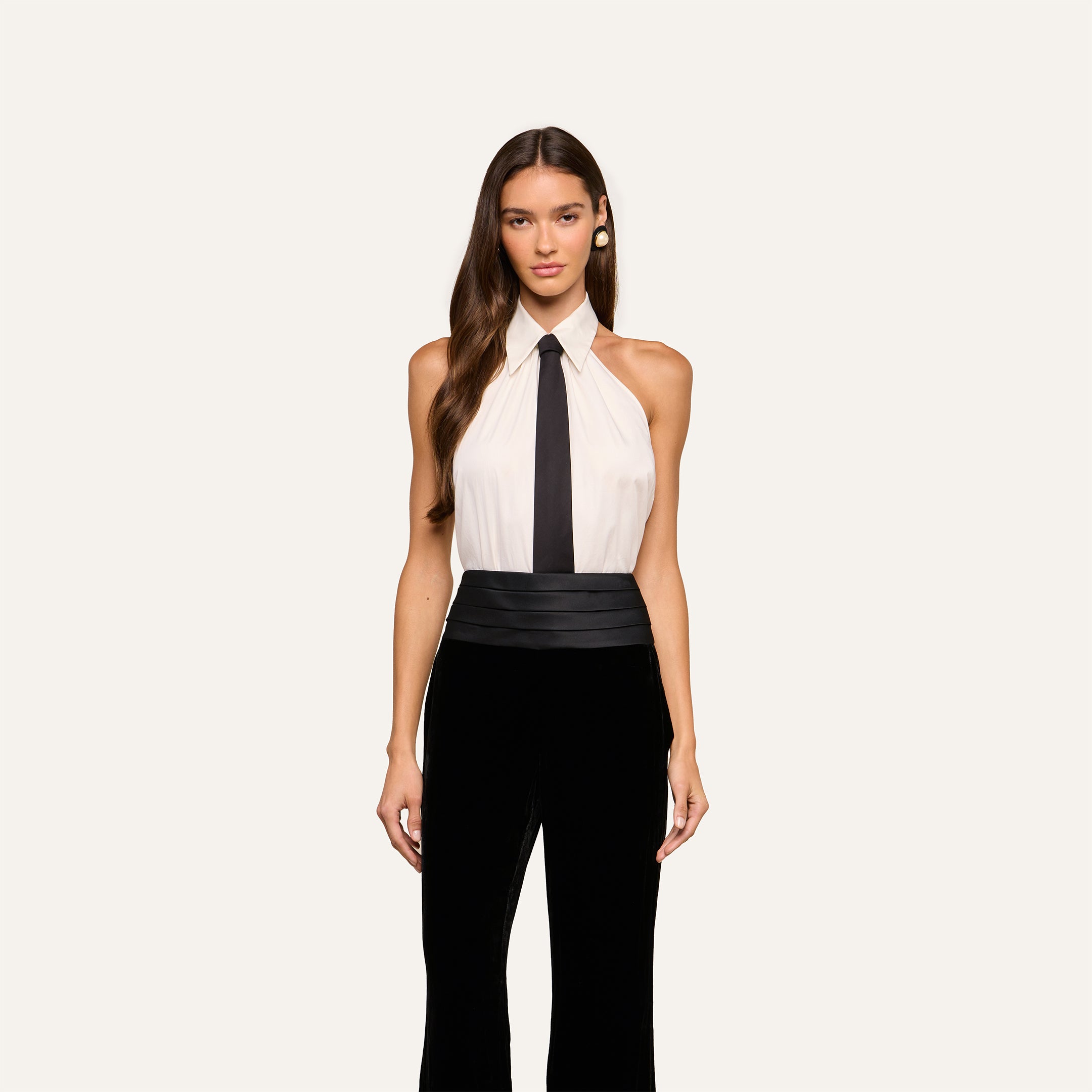 A woman with long brown hair wears the Wren Neck-Tie Top— a sleeveless, halter black and white button-down piece featuring a black tie detail—paired with wide-leg pants, standing against a plain light background.