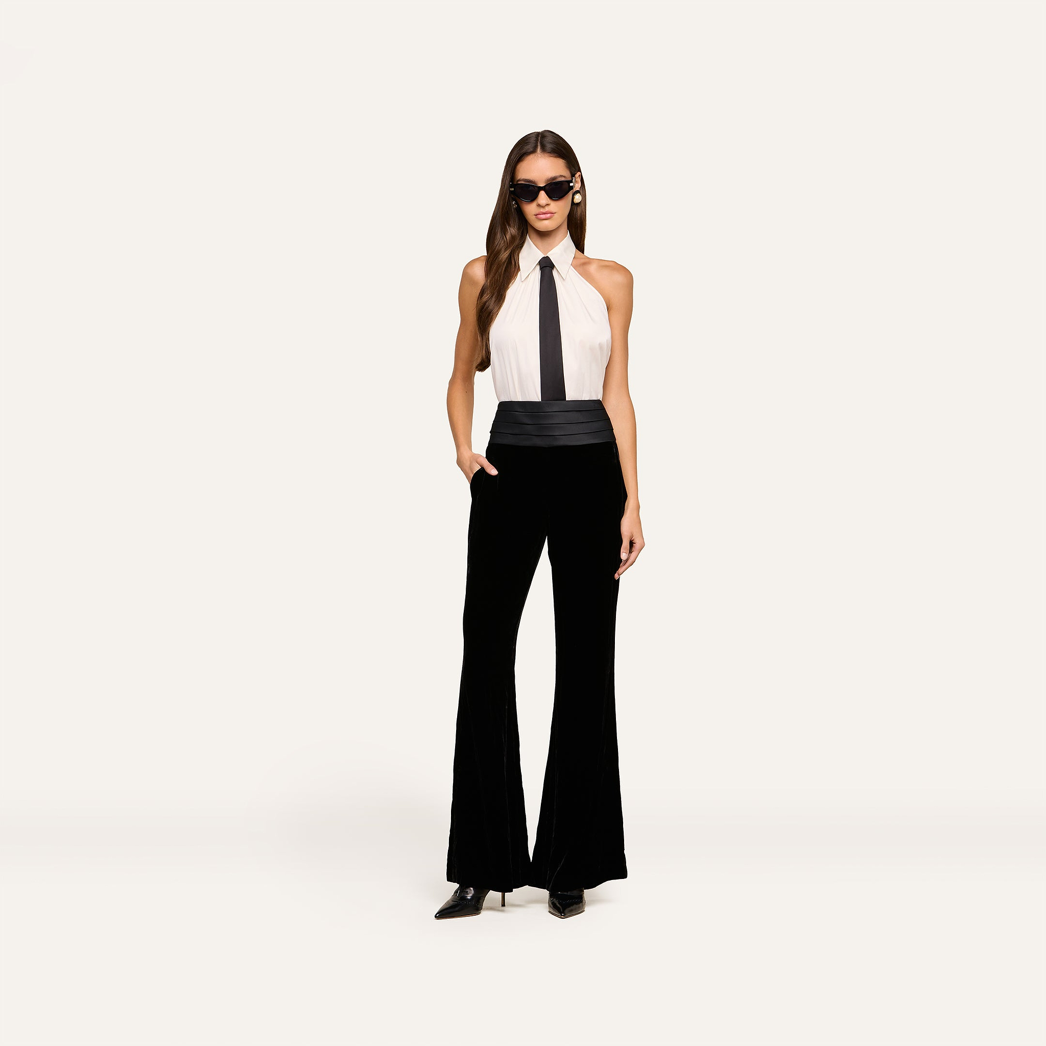 A woman wearing sunglasses and the Wren Neck-Tie Top—a sleeveless white halter button-down with a black necktie—paired with high-waisted black flared pants stands confidently against a plain light background.