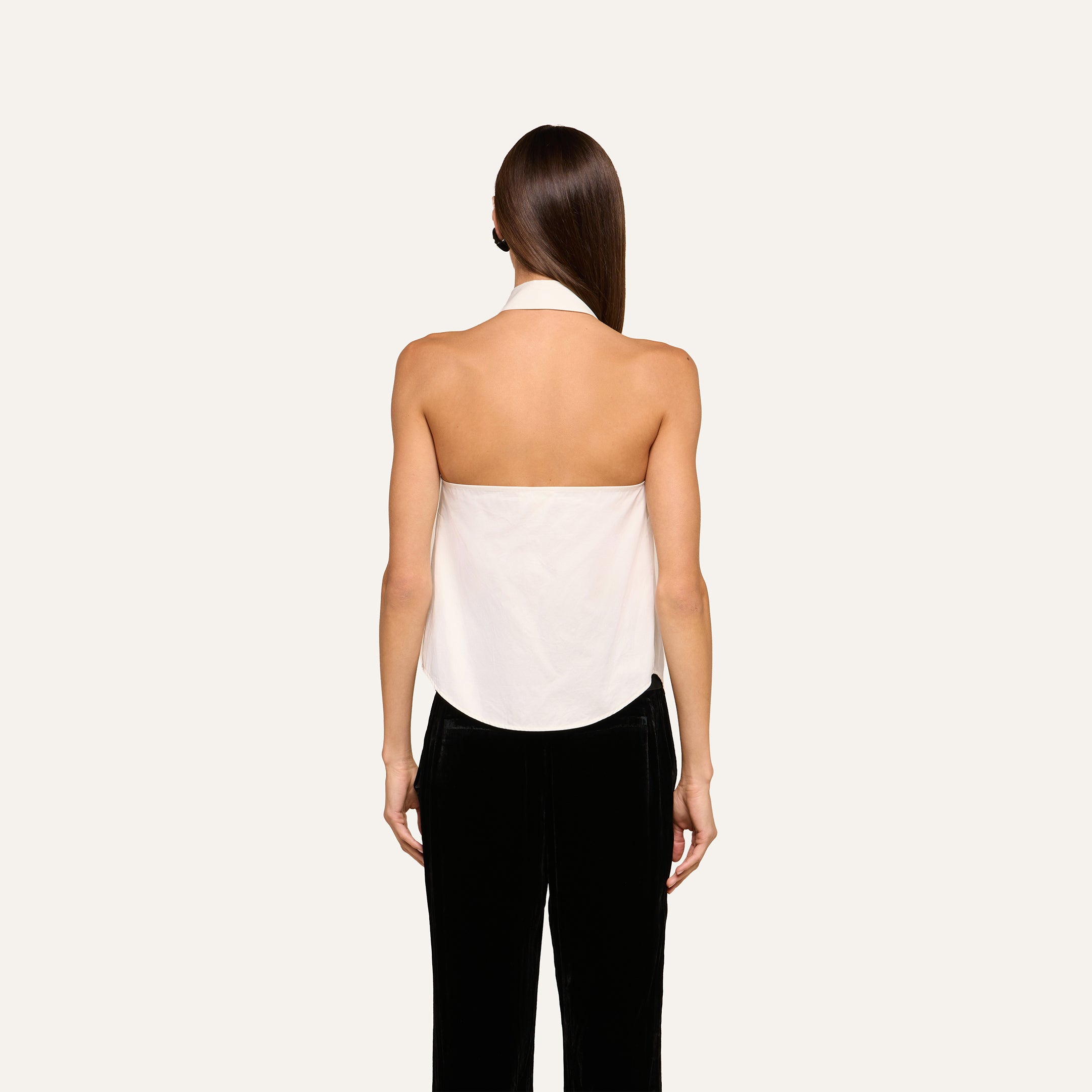 A woman stands facing away against a plain light background, wearing the Wren Neck-Tie Top—a white halter button-down style—paired with black pants. Her straight brown hair is down and her arms are at her sides.