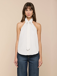 Una donna dai lunghi capelli castani indossa il Wren Poplin Top, un top bianco senza maniche con dettaglio di cravatta in morbido popeline sateen, abbinato a jeans blu scuro, su un semplice sfondo beige.