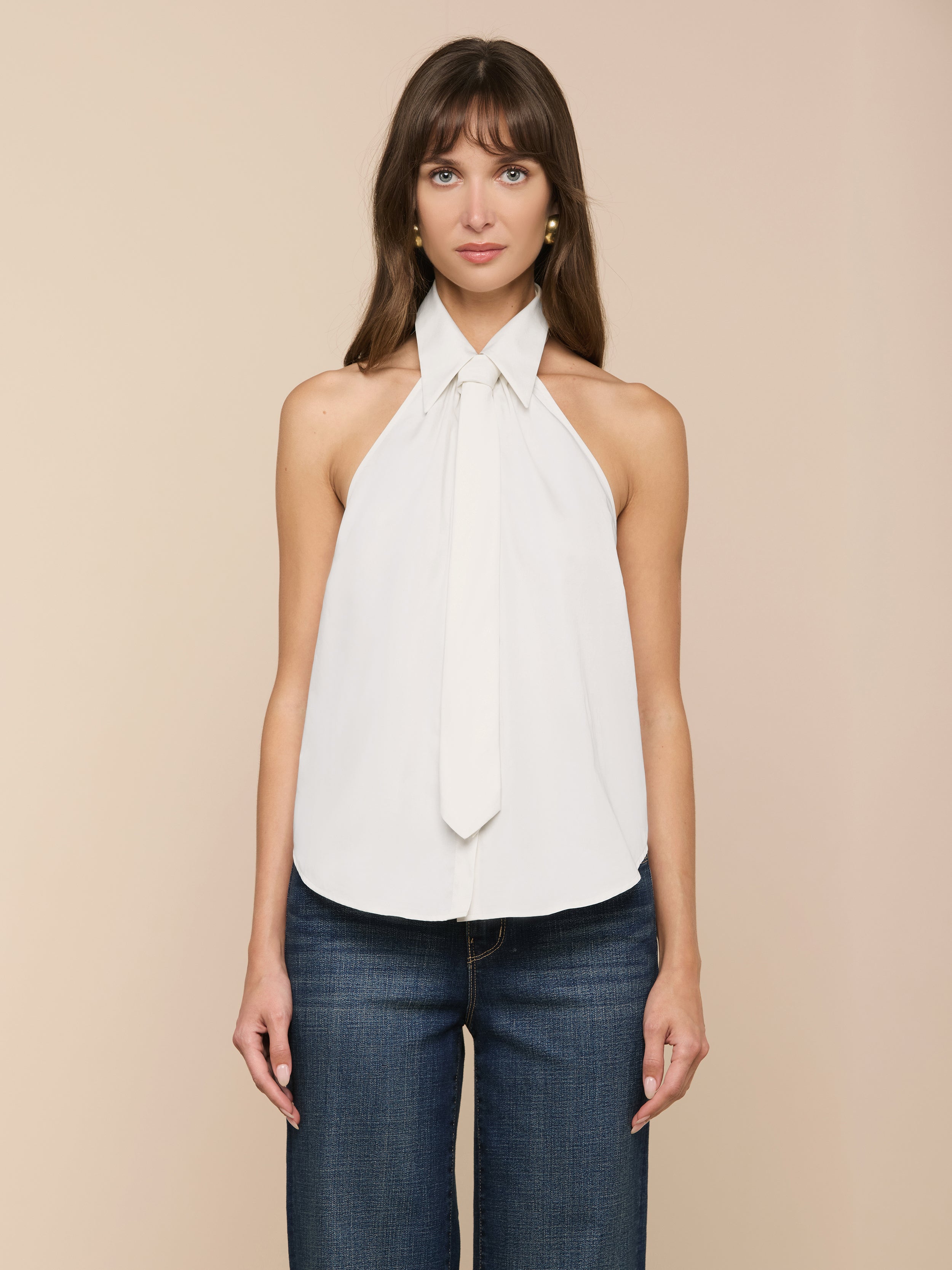 Una donna dai lunghi capelli castani indossa il Wren Poplin Top, un top bianco senza maniche con dettaglio di cravatta in morbido popeline sateen, abbinato a jeans blu scuro, su un semplice sfondo beige.