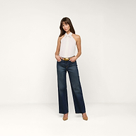Una donna in piedi su uno sfondo beige indossa il top Wren Poplin bianco senza maniche con collo ampio, jeans blu scuro a gamba larga, cintura e tacchi marroni. Ha le mani in tasca e unespressione neutra.