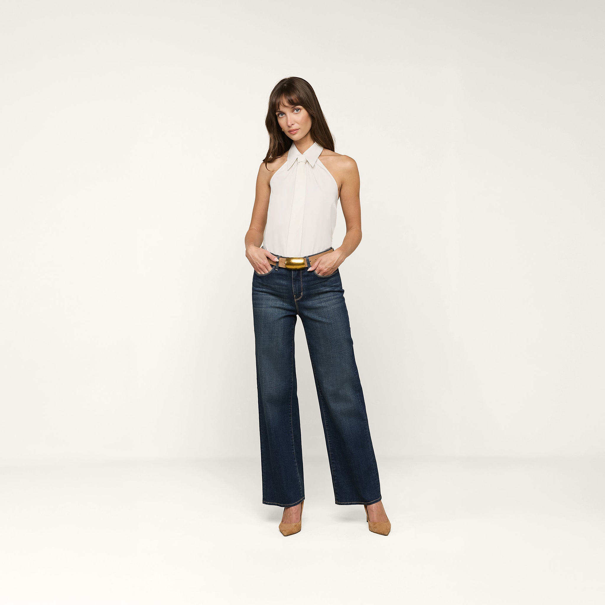 Una donna in piedi su uno sfondo beige indossa il top Wren Poplin bianco senza maniche con collo ampio, jeans blu scuro a gamba larga, cintura e tacchi marroni. Ha le mani in tasca e unespressione neutra.