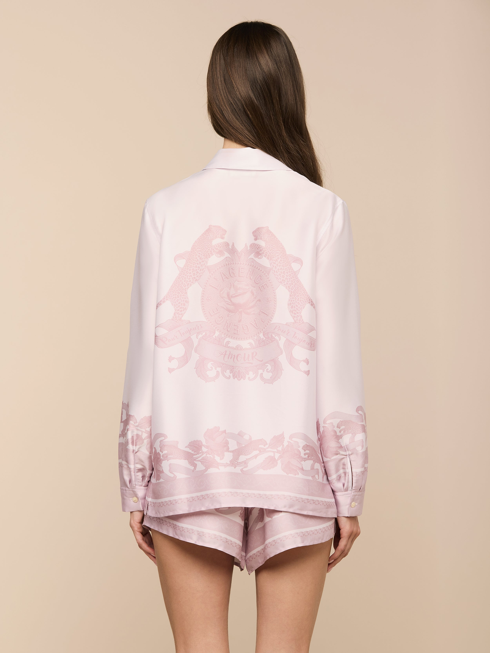 Wearing the Judy Blouse, a woman with long brown hair stands with her back to the camera against a plain, light background. The light pink button-down pajama set features decorative patterns and a crest on the back.