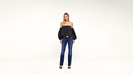 Woman stands against a plain white background wearing a black off-the-shoulder top with puffy sleeves, blue jeans, black heels, and a large belt, facing forward with arms at her sides.