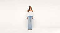 A woman stands facing forward against a plain white background, wearing a white off-the-shoulder top and light blue jeans.