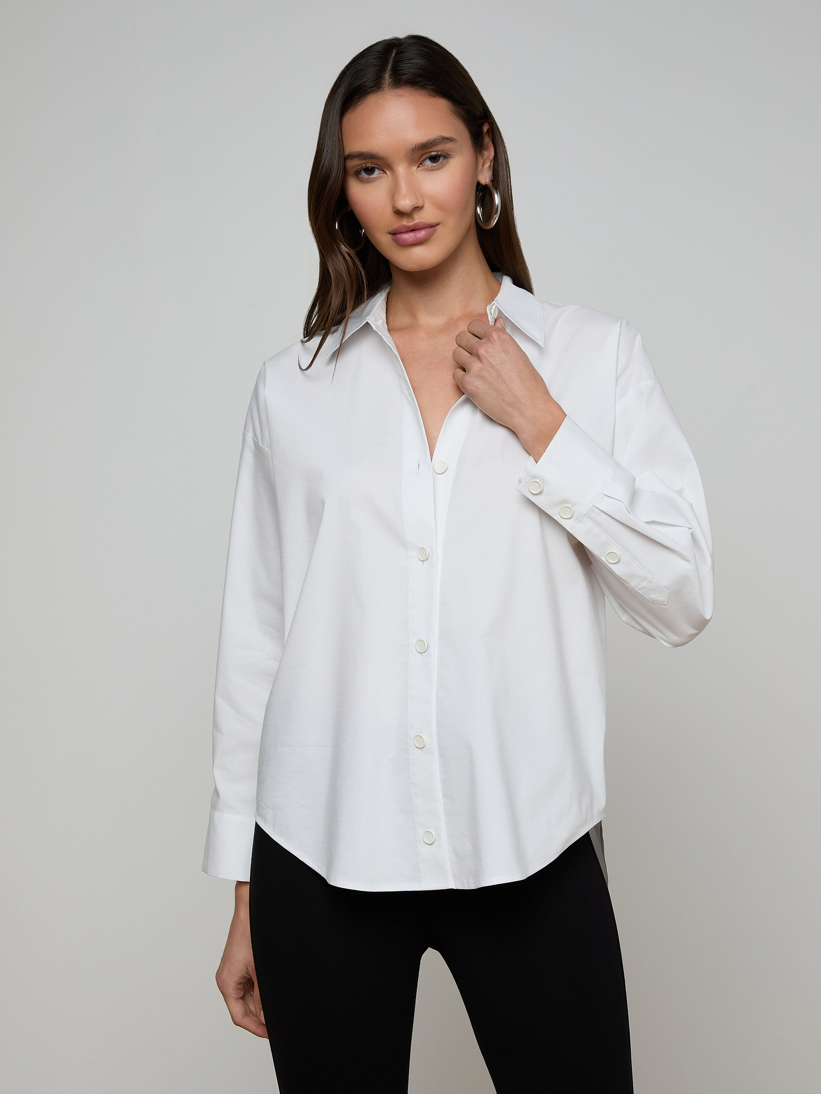 A woman wears the Rylie Poplin Shirt—a relaxed fit, white button-down crafted from organic cotton—paired with black pants. She stands against a plain light background, holding the shirt’s collar with one hand and looking at the camera.