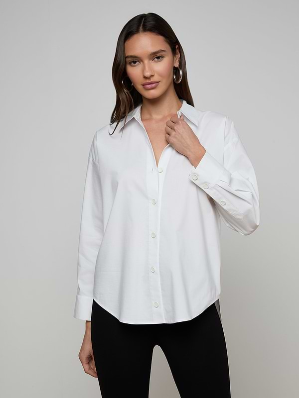 A woman wears the Rylie Poplin Shirt—a relaxed fit, white button-down crafted from organic cotton—paired with black pants. She stands against a plain light background, holding the shirt’s collar with one hand and looking at the camera.