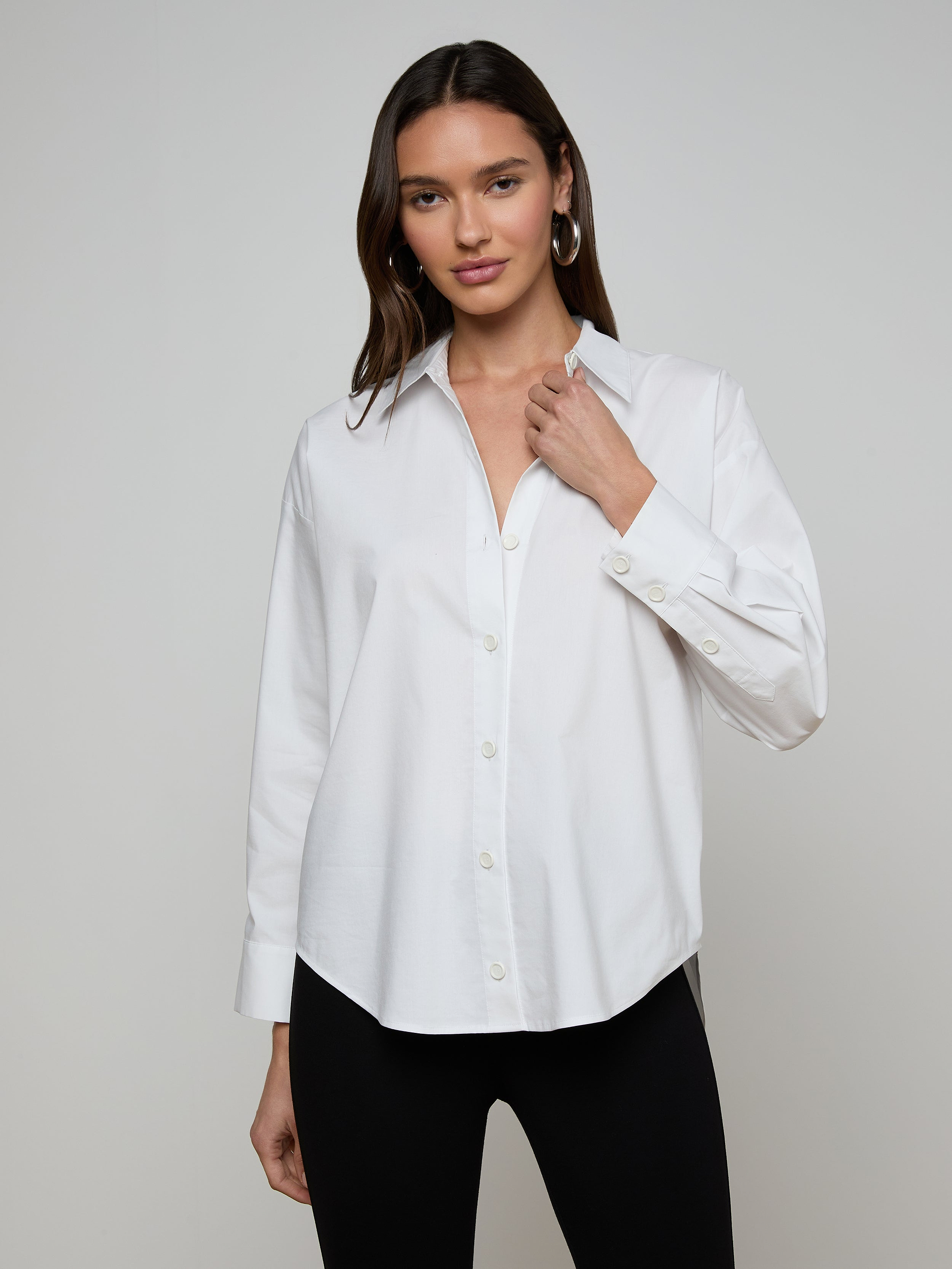 A woman wears the Rylie Poplin Shirt—a relaxed fit, white button-down crafted from organic cotton—paired with black pants. She stands against a plain light background, holding the shirt’s collar with one hand and looking at the camera.