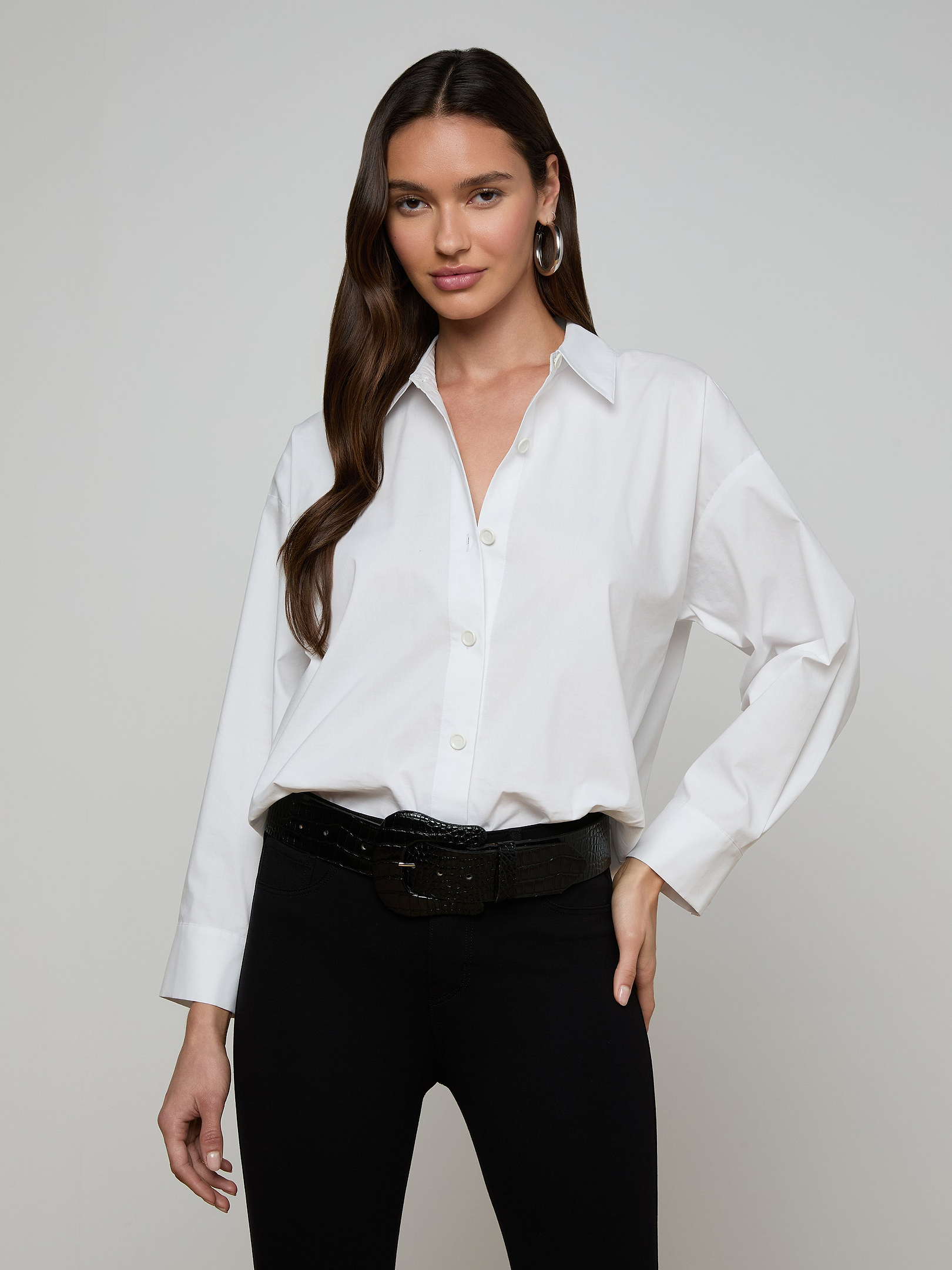 Woman with long brown hair wearing the Rylie Poplin Shirt—a white button-down made from organic cotton—paired with black pants, a black belt, and hoop earrings, standing with one hand on her hip against a plain light background.