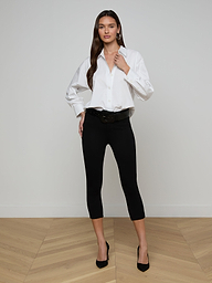 A woman stands against a plain white background, wearing the Rylie Poplin Shirt—a relaxed fit white button-down made from organic cotton. She pairs it with black cropped pants and black heels, her long brown hair cascading down as she poses with a confident expression.