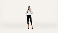 A woman stands against a plain white background, wearing a white blouse, black cropped pants, a black belt, and black heels. She has long brown hair and poses with one hand on her waist and the other touching her blouse.