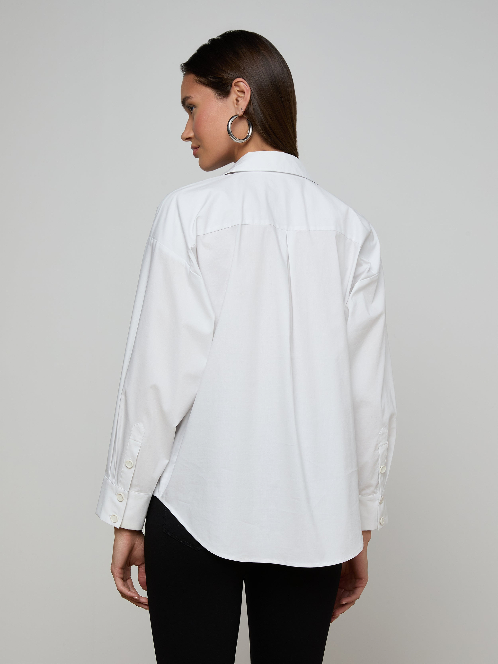 A woman with straight brown hair stands with her back to the camera against a plain light background, wearing the Rylie Poplin Shirt—a relaxed fit, white button-down made from organic cotton—paired with black pants. She accessorizes with silver hoop earrings, and her left sleeve is partially rolled up.