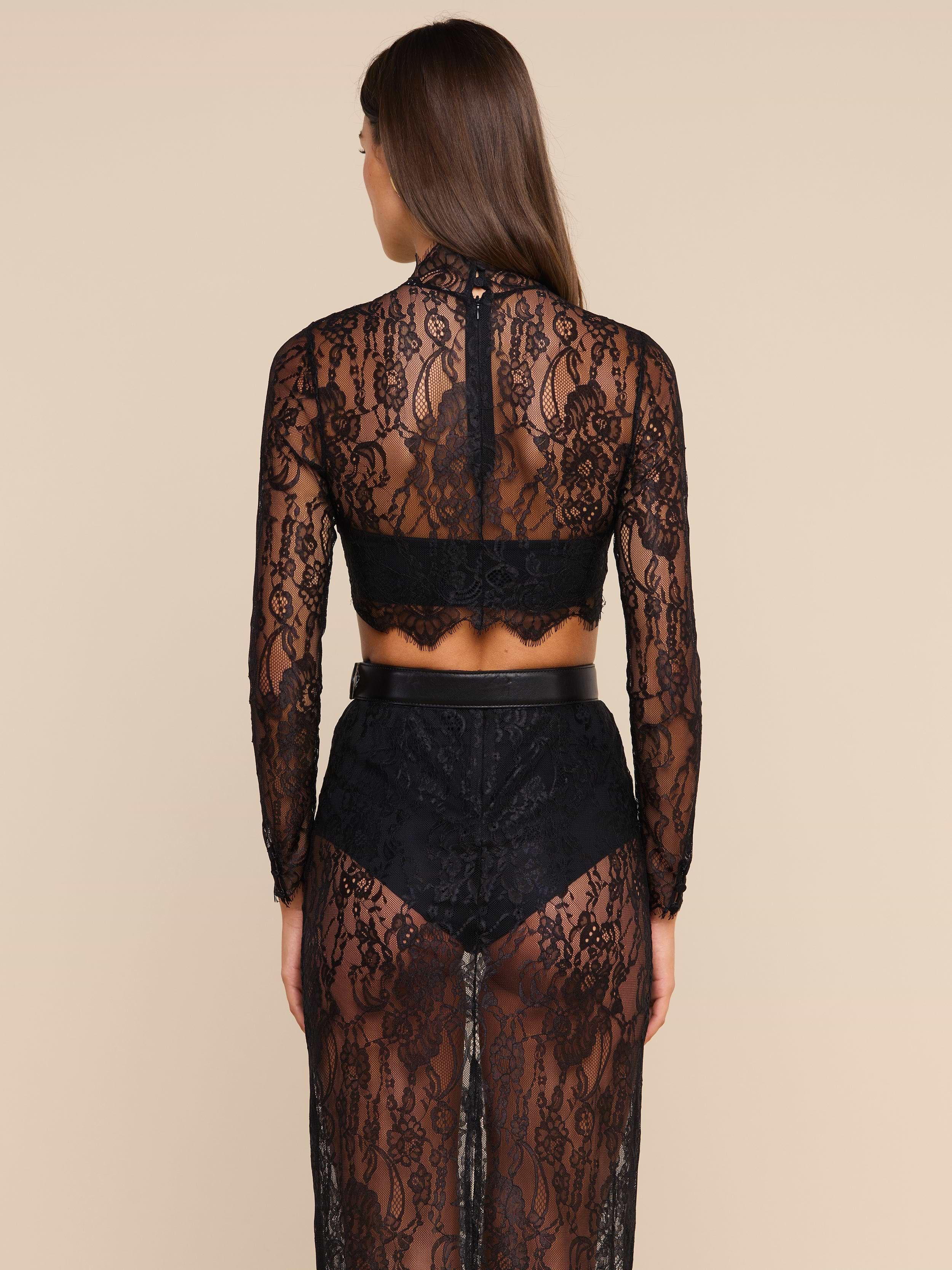A woman with long brown hair stands facing away, wearing the Henriette Cropped Lace Top with long sleeves and a matching high-waisted skirt, revealing black undergarments underneath. The outfit is detailed with delicate scalloped hems, set against a plain light-colored background.