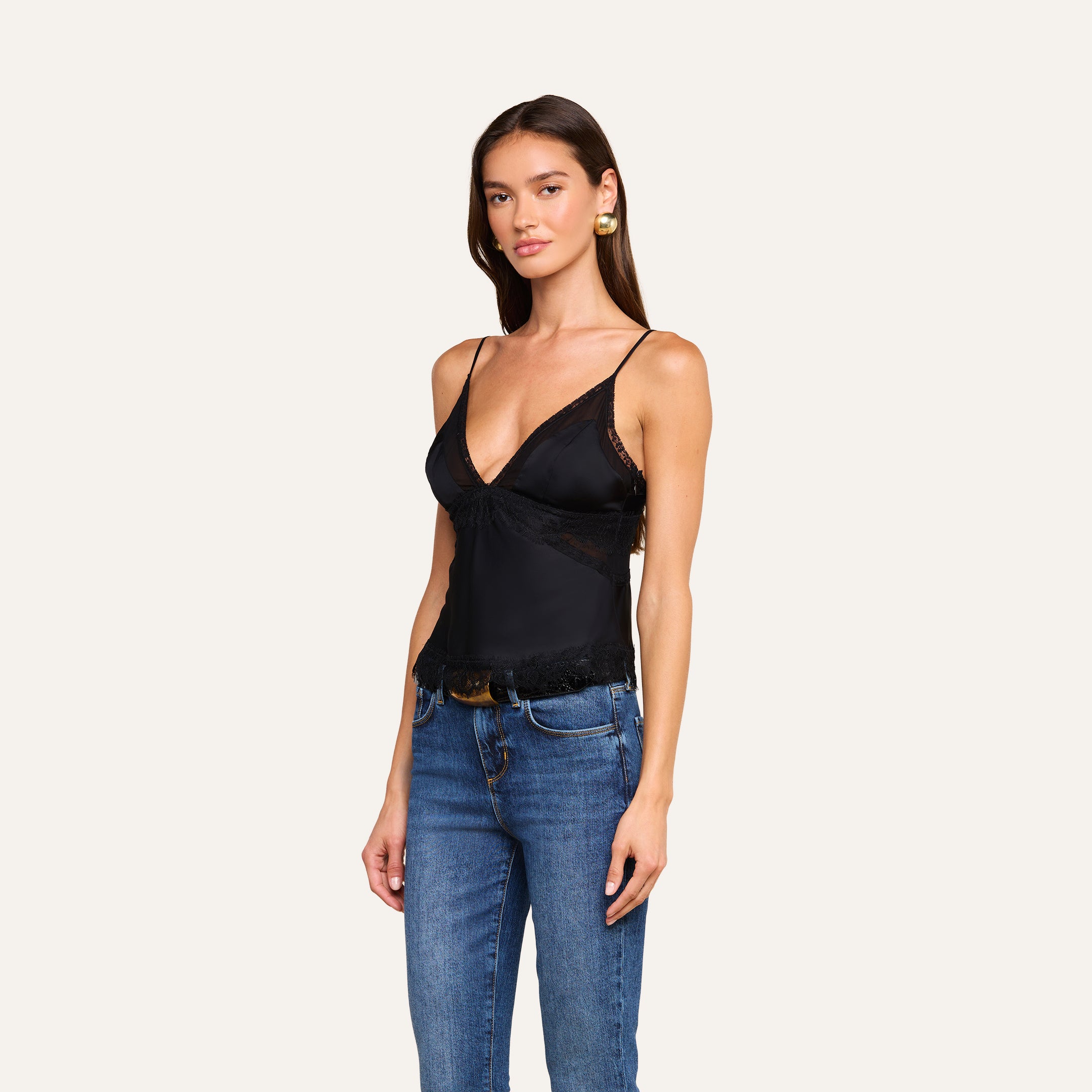 A woman with long brown hair wears the Elka Satin Lace Camisole—a sleeveless black satin top with lace trim—paired with blue jeans. She stands against a plain light background, looking at the camera with a slight smile.