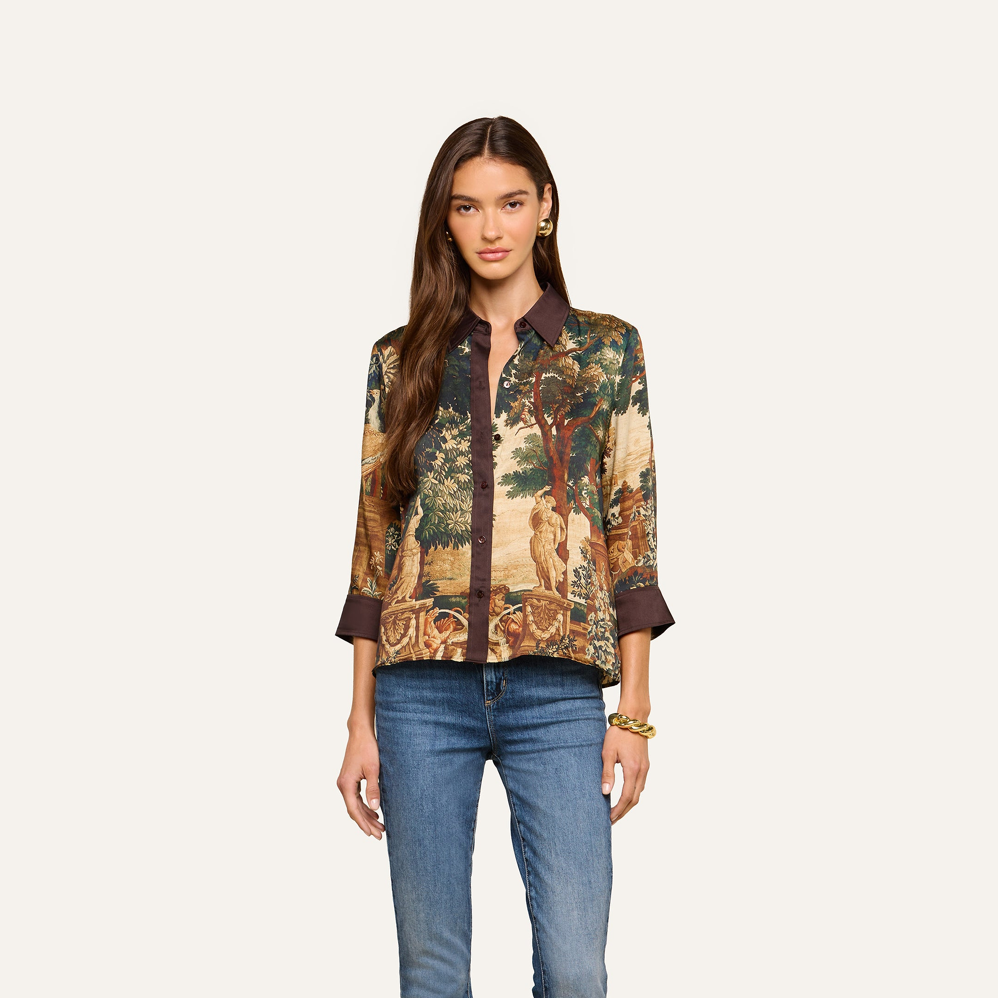 A woman with long brown hair is wearing the Eulalie Blouse—a tapestry print top featuring a dark collar and cuffs—paired with blue jeans and a gold bracelet as she stands against a plain light background.
