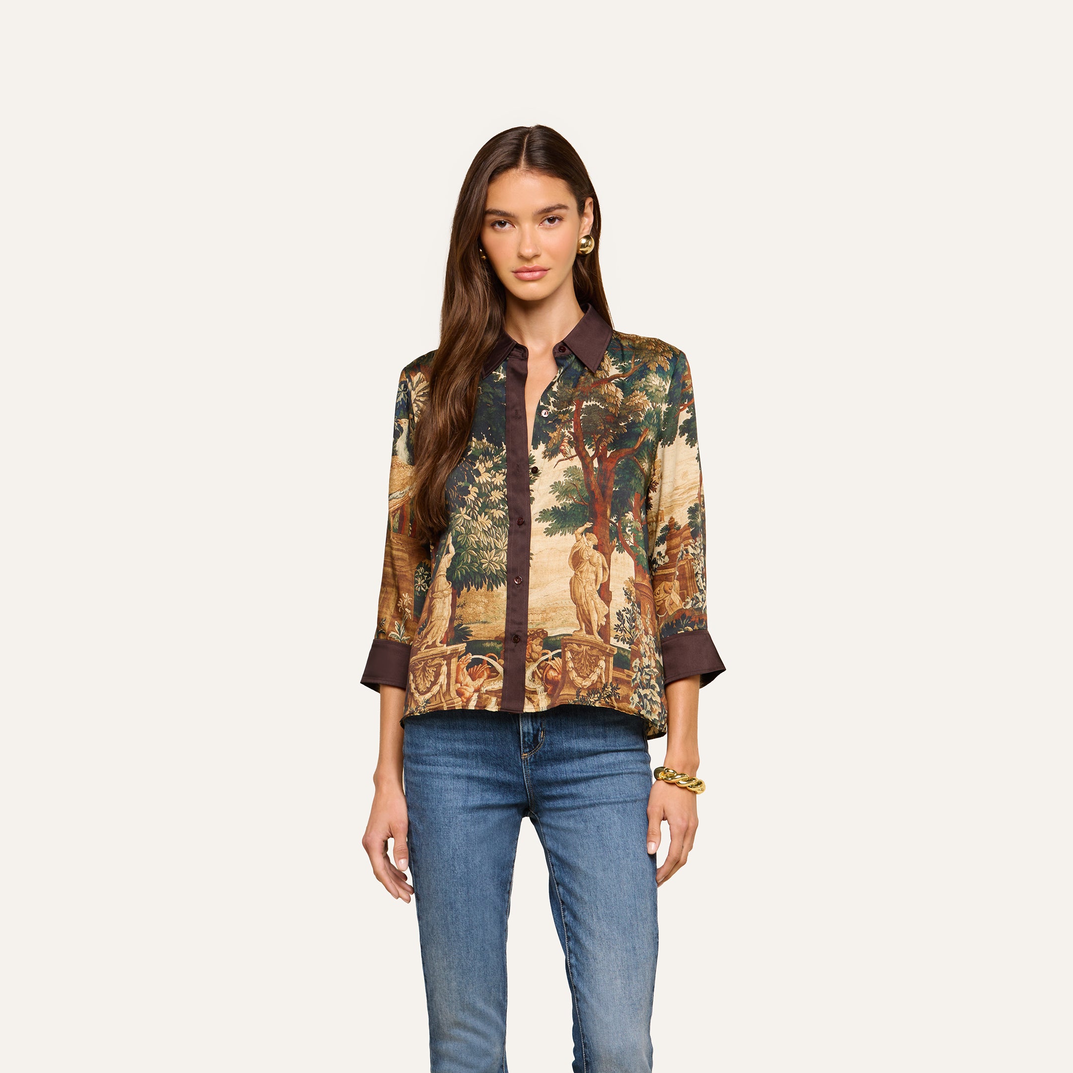 A woman with long brown hair is wearing the Eulalie Blouse—a tapestry print top featuring a dark collar and cuffs—paired with blue jeans and a gold bracelet as she stands against a plain light background.