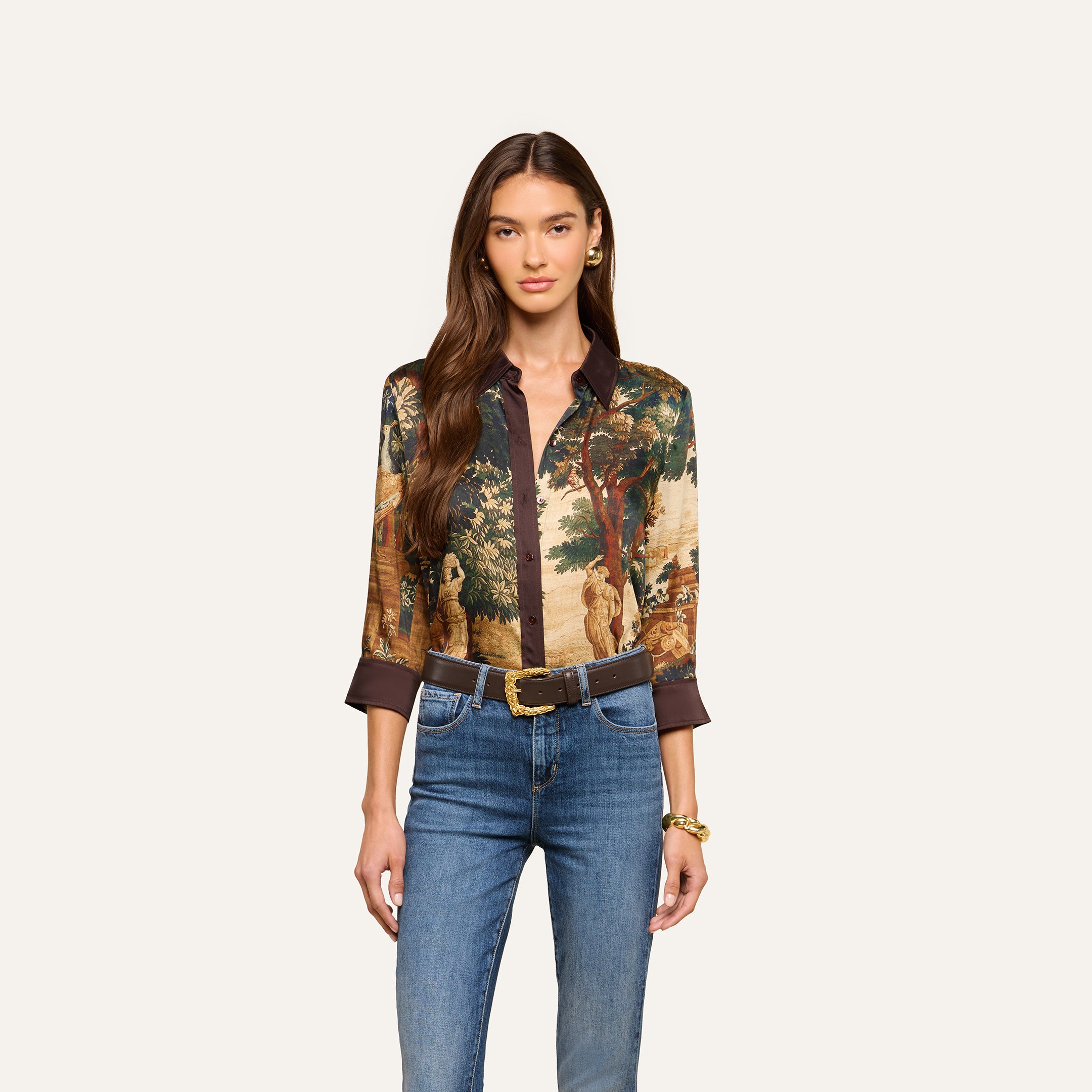 Woman with long brown hair wearing the Eulalie Blouse, featuring a tapestry-inspired scenic print, styled with blue jeans, a belt, and gold jewelry, standing against a plain light background.