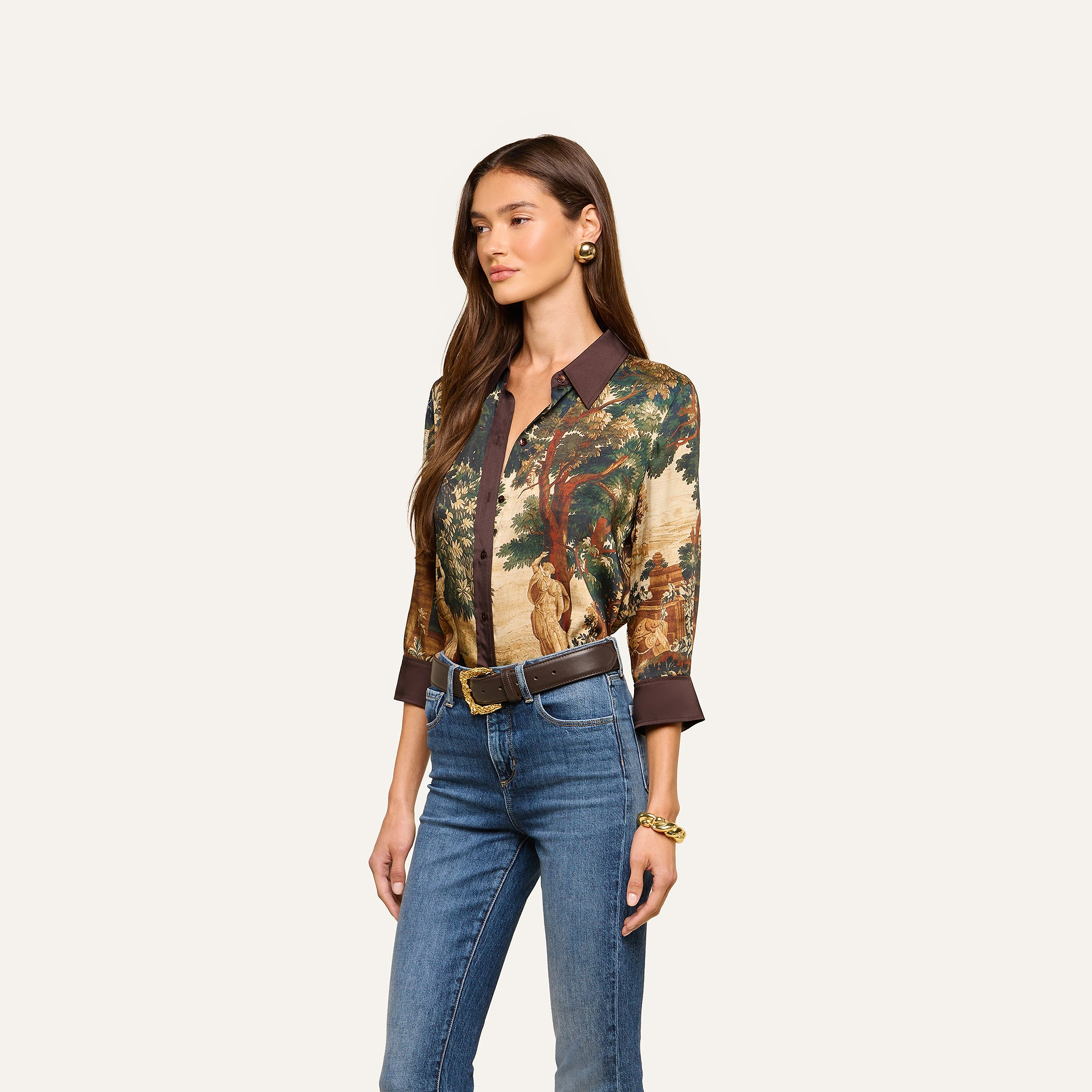 A woman wearing the Eulalie Blouse—a tapestry print blouse featuring a nature scene—paired with blue jeans, a dark belt, gold earrings, and a gold bracelet, stands against a plain light background.