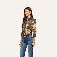 A woman wearing the Eulalie Blouse—a tapestry print long-sleeve blouse featuring a nature scene—paired with blue jeans, a belt, and gold jewelry, stands against a plain light background.