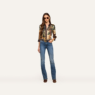 A woman stands against a plain light background, looking confidently at the camera with her hands in her pockets. She is wearing sunglasses, blue jeans, brown shoes, and the Eulalie Blouse.
