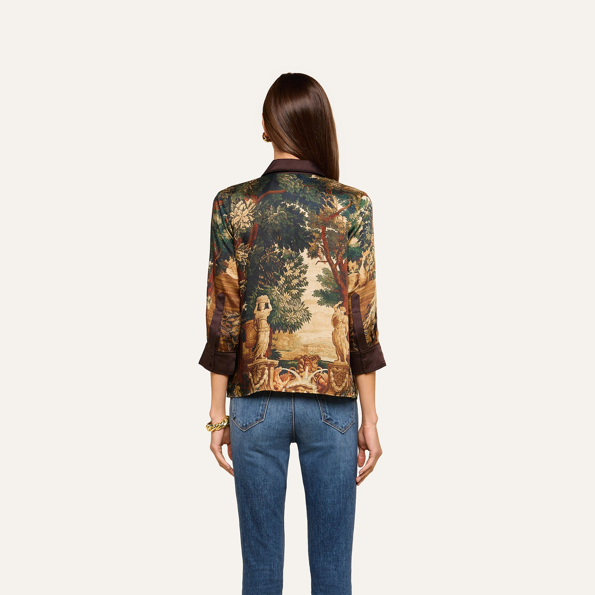 A person with long brown hair, wearing the Eulalie Blouse with a tapestry-style nature scene print and blue jeans, stands facing away from the camera against a plain background.