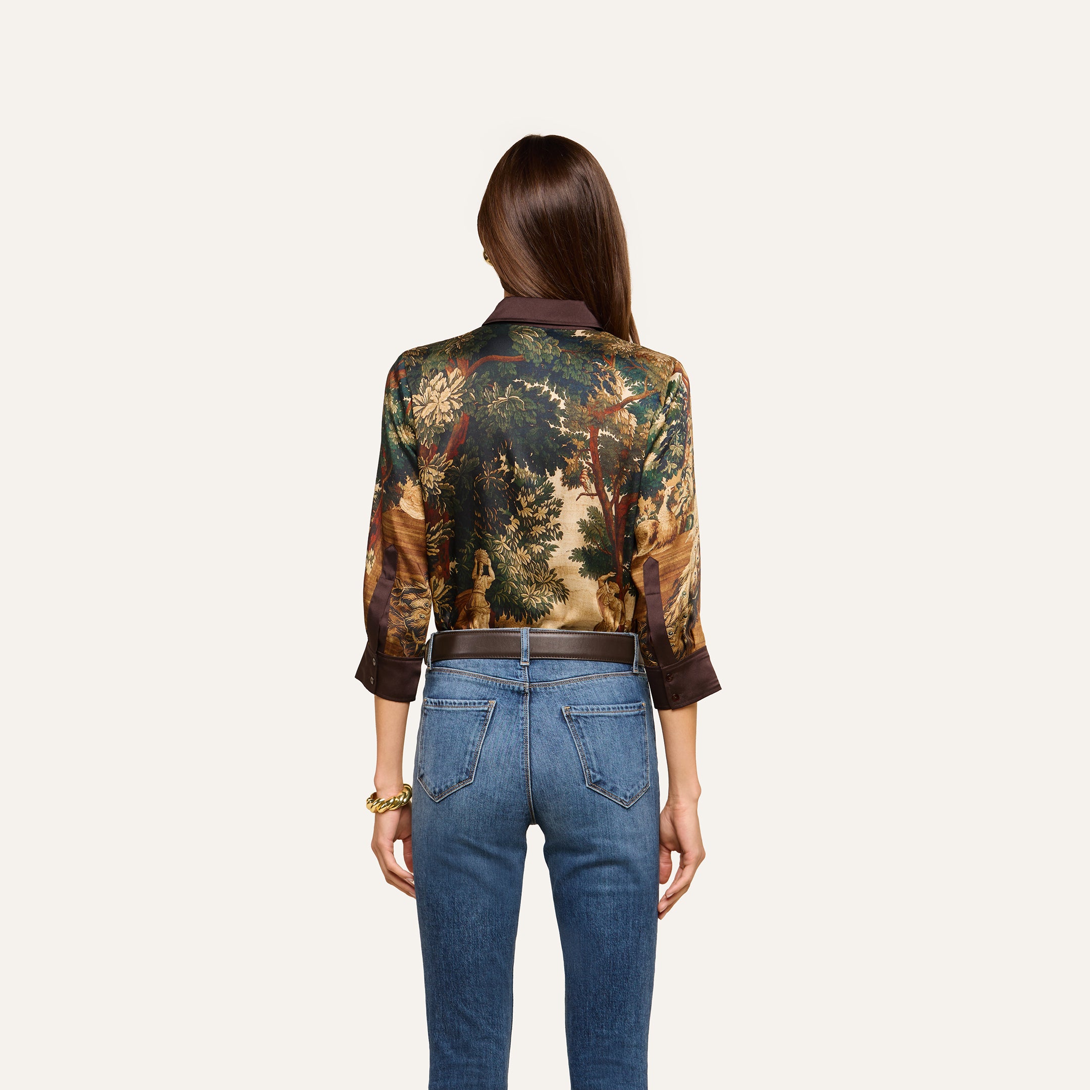 A woman with long brown hair, seen from behind, wears the Eulalie Blouse, featuring a tapestry print with a nature scene, paired with blue jeans. She stands against a plain light background.