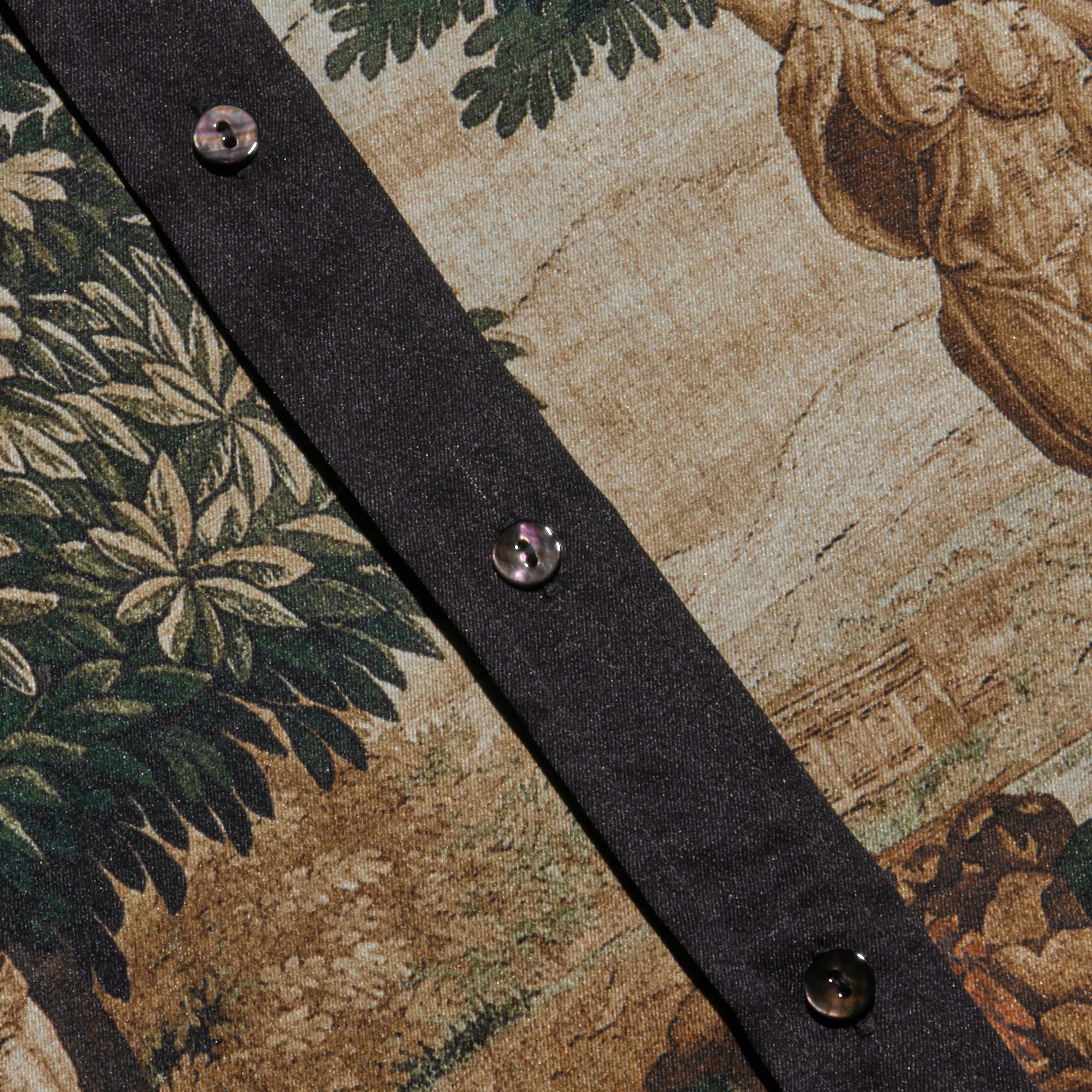 A close-up of the Eulalie Blouse showcases its fitted silhouette and scenic printed pattern featuring trees, foliage, and a building. A dark fabric strip adorned with three black buttons runs diagonally across the blouse.
