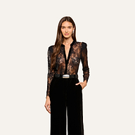 A woman with long brown hair wearing the black Dorothea Velvet Lace Blouse and high-waisted black pants, one hand in her pocket, stands against a plain light background.