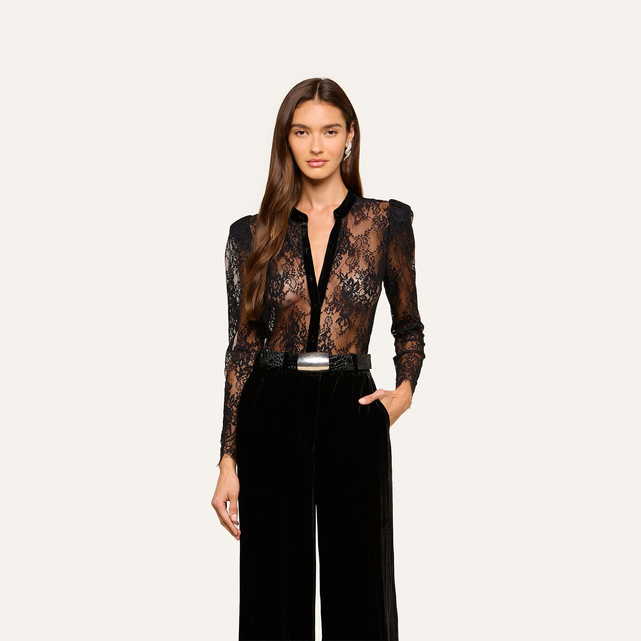 A woman with long brown hair wearing the black Dorothea Velvet Lace Blouse and high-waisted black pants, one hand in her pocket, stands against a plain light background.