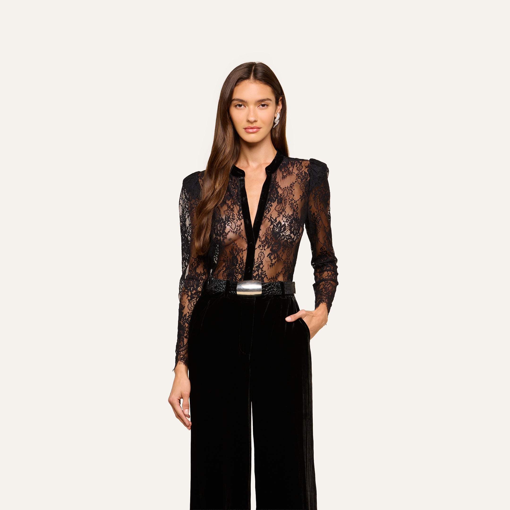 A woman with long brown hair wearing the black Dorothea Velvet Lace Blouse and high-waisted black pants, one hand in her pocket, stands against a plain light background.