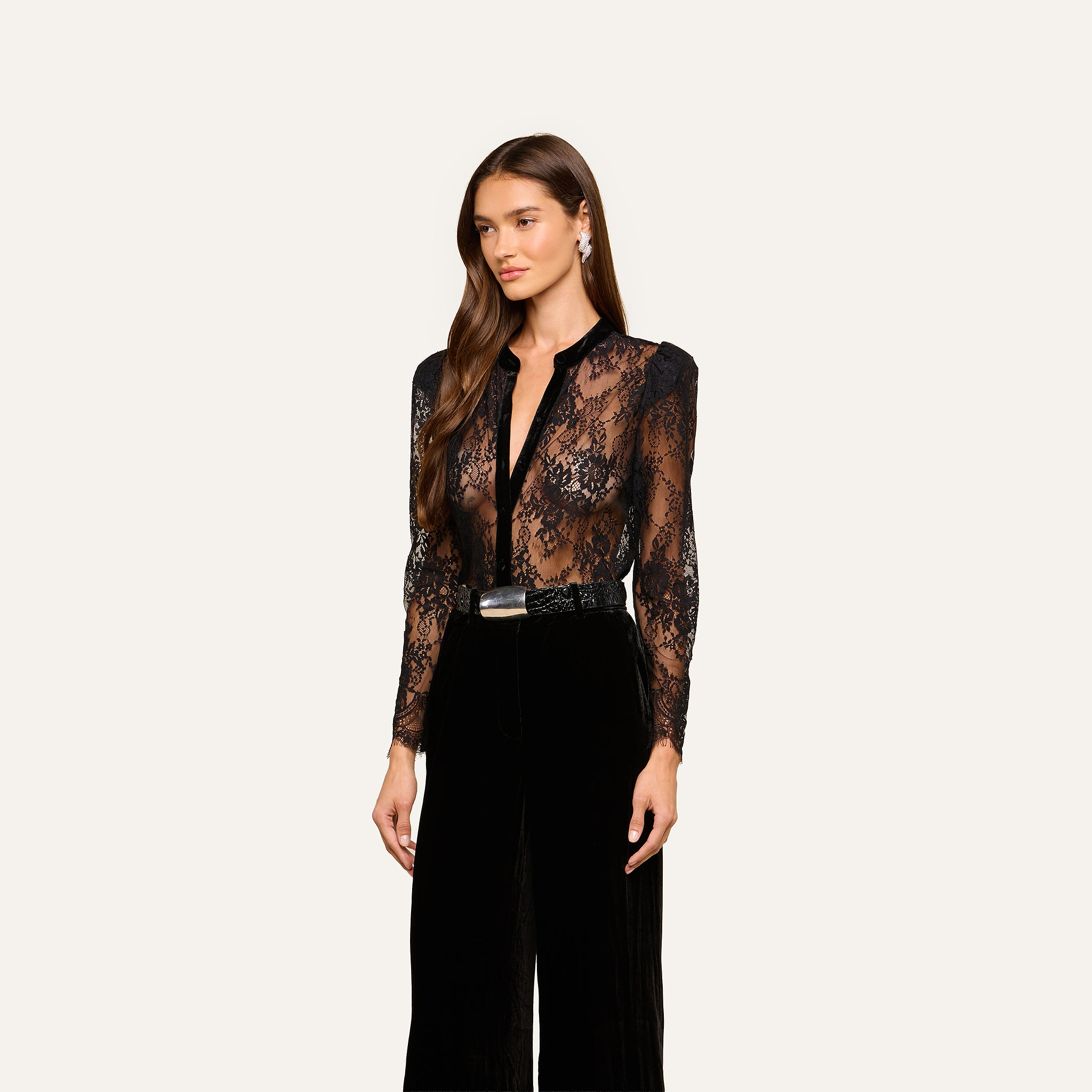 A woman with long brown hair wears the Dorothea Velvet Lace Blouse, paired with black high-waisted pants, a black belt with a silver buckle, and drop earrings, standing against a plain light background.