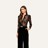 A woman wearing the Dorothea Velvet Lace Blouse—a black long-sleeve top—paired with high-waisted black pants stands against a light background, looking to the side with her hair down.