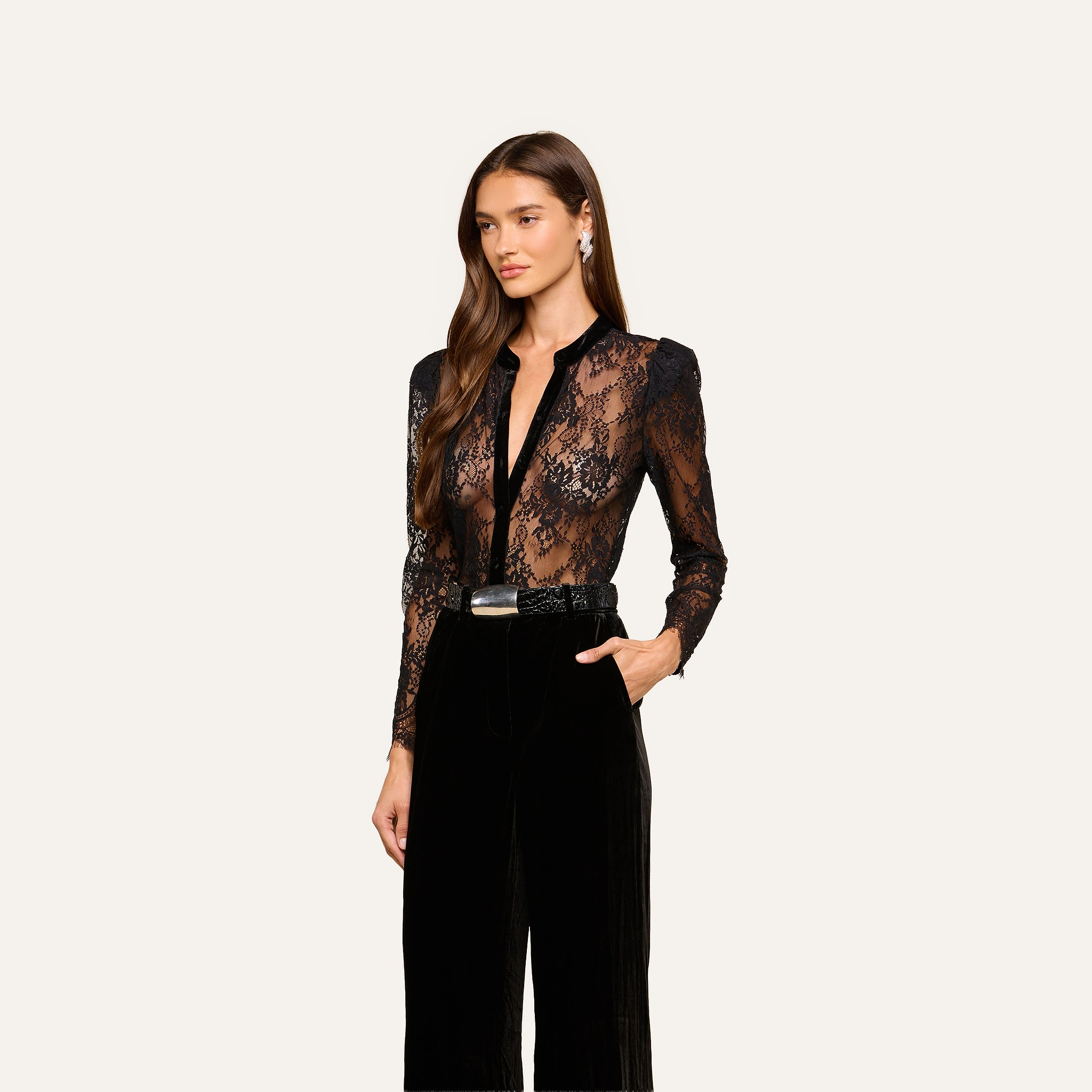 A woman wearing the Dorothea Velvet Lace Blouse—a black long-sleeve top—paired with high-waisted black pants stands against a light background, looking to the side with her hair down.