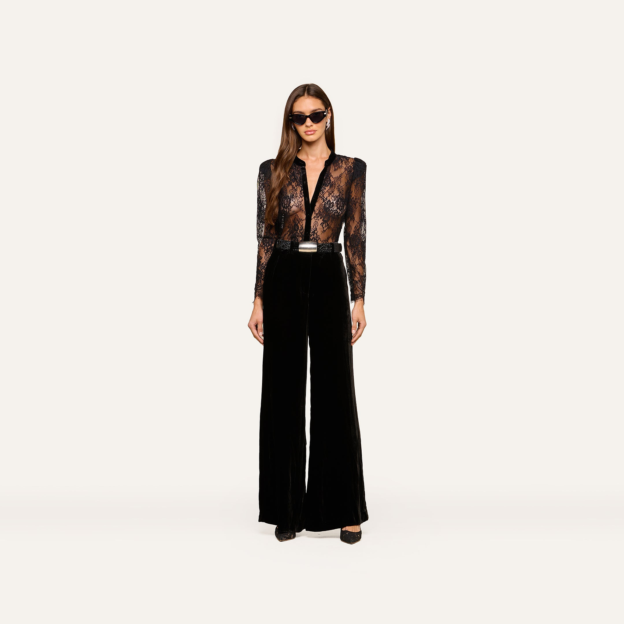A woman wearing sunglasses, the Dorothea Velvet Lace Blouse—featuring sheer Chantilly lace with velvet seams—and high-waisted black wide-leg pants stands facing forward against a plain white background.