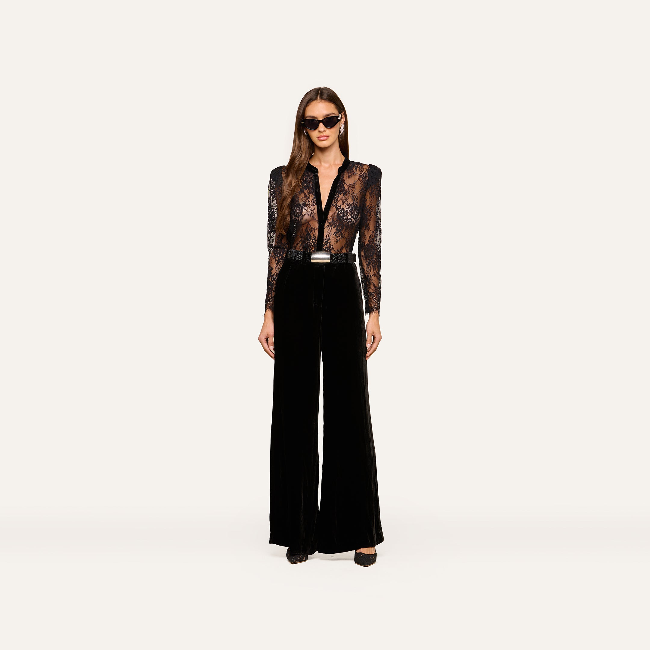 A woman wearing sunglasses, the Dorothea Velvet Lace Blouse—featuring sheer Chantilly lace with velvet seams—and high-waisted black wide-leg pants stands facing forward against a plain white background.
