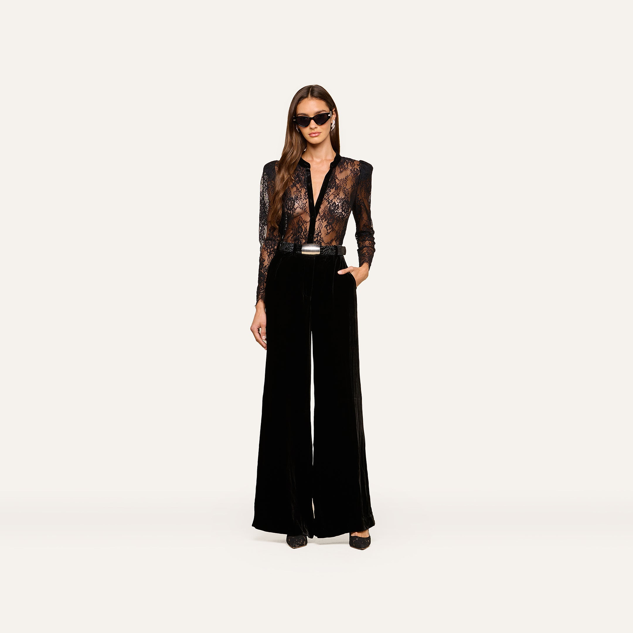 Wearing the Dorothea Velvet Lace Blouse, a woman in sunglasses stands confidently with one hand in her pocket, showcasing its sheer Chantilly lace long sleeves tucked into high-waisted, wide-leg black velvet pants with elegant velvet seams and finished with black shoes.